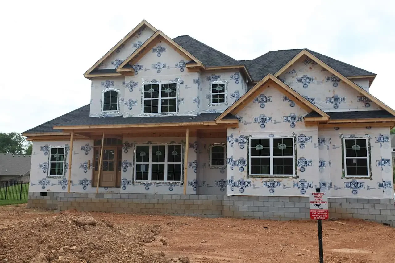 Two-story house under construction with wrap-around windows, a covered front porch, and a shingled roof. The exterior is unfinished with visible house wrap and bricks at the foundation.