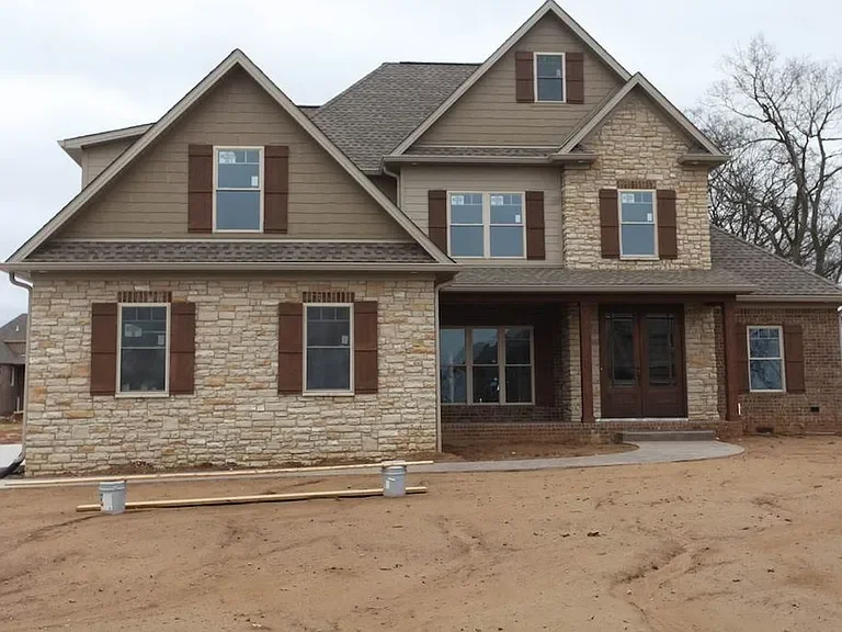 New two-story house with stone and siding exterior, multiple windows with brown shutters, and a front porch with steps, surrounded by bare soil ground.