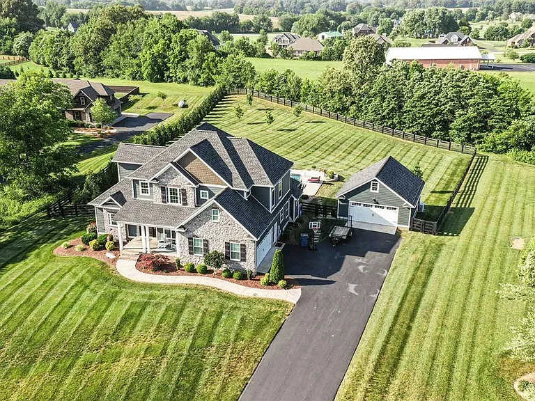 An aerial view of a large two-story house with a gray exterior and dark roof, surrounded by a well-maintained lawn, a long driveway, and a detached garage. There is a backyard with a small deck and a fenced area, set in a rural area with open fields 