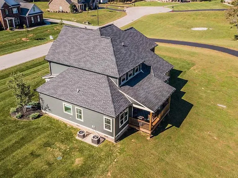 An aerial view of a two-story house with a dark gray roof and light gray exterior in a suburban neighborhood. The house has a small backyard deck, several windows, and is surrounded by a large grassy lawn. Other houses and a small golf green are visi