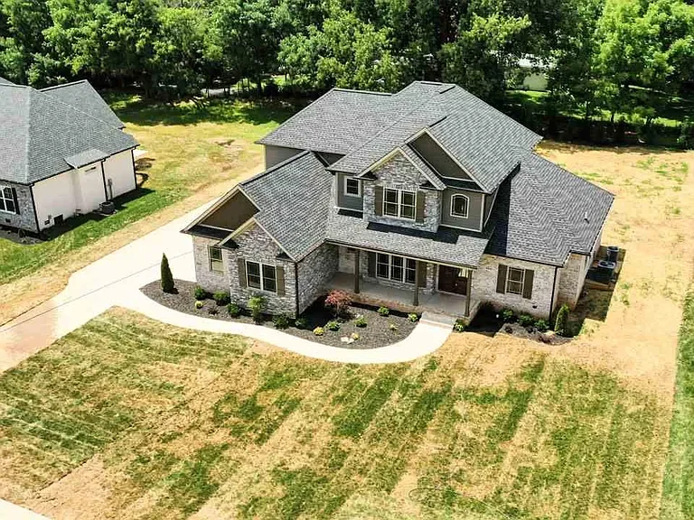 An aerial view of a large, modern house with gray roofing and stone exterior, surrounded by a partially landscaped yard with small bushes and trees, and neighboring houses in the background.