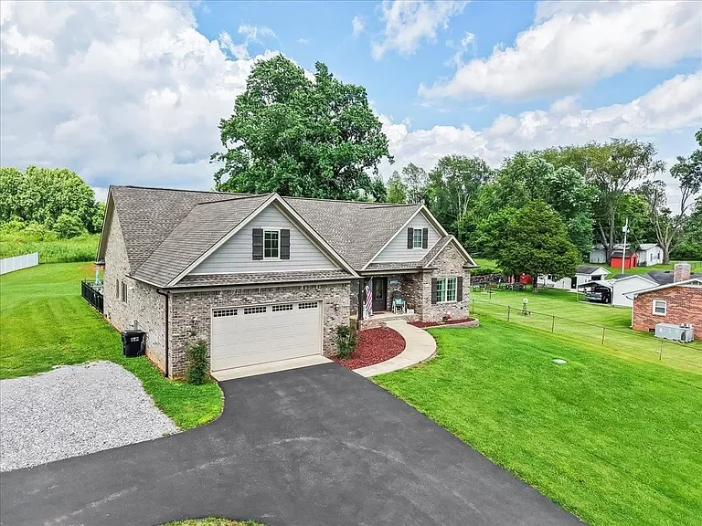 A suburban house with a brick and siding exterior, a gray roof, and a white garage door. The house has a curved walkway, green lawn, and scattered trees in the background under a partly cloudy sky.