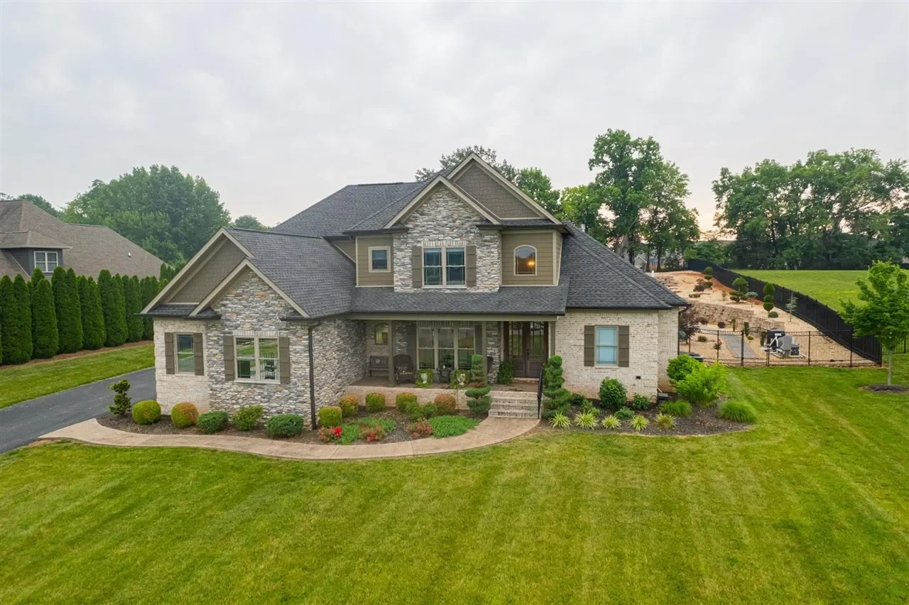 Front view of a large, modern house with stone and siding exterior, surrounded by a lush green lawn and landscaped garden.