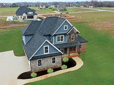 A two-story house with gray roofing, brick and white siding, surrounded by a lawn with small bushes.
