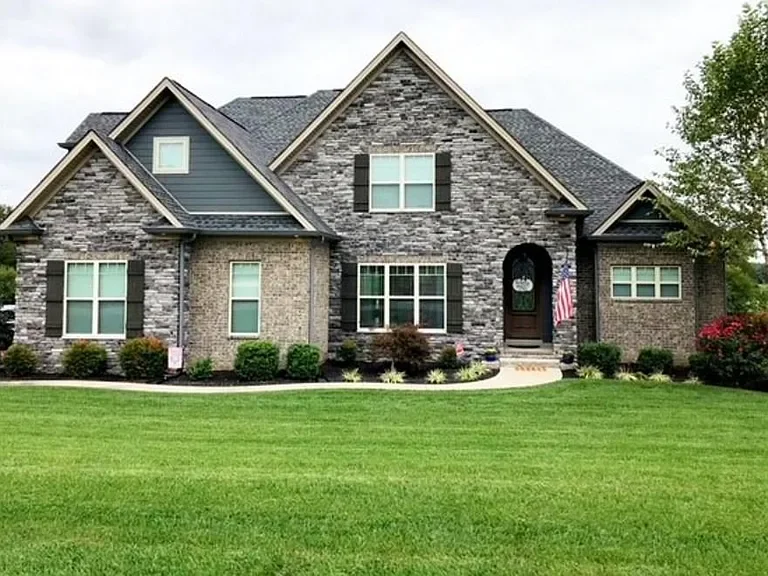 Large brick house with a stone facade, four front-facing windows, black shutters, and a landscaped front yard.