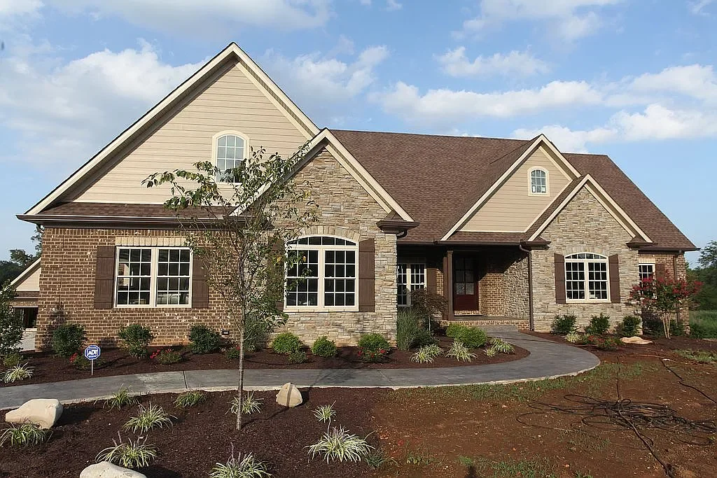 Single-story house with brick and stone exterior and a brown shingle roof, front porch, several large windows with shutters, and a landscaped front yard with young plants and a tree.