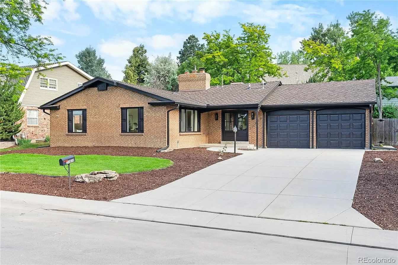 A single-story brick house with a two-car garage, black garage doors, and black window trims. The front yard is landscaped with green grass and mulch, with a small mailbox near the driveway and trees surrounding the house. The sky is partly cloudy.