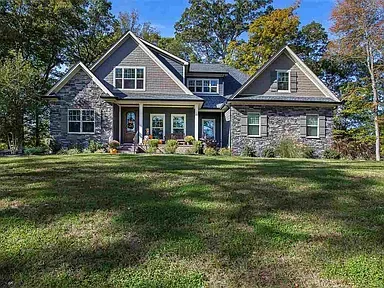 A large two-story house with gray stone and siding exterior, multiple windows, and a front porch set in a grassy yard with trees in the background.