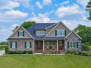 Large modern house with brick and siding exterior, front porch with chairs, surrounded by a well-maintained lawn and trees under a partly cloudy sky.
