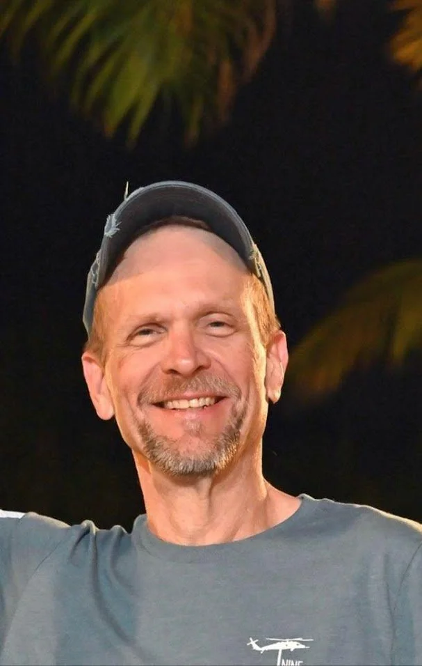 A smiling man wearing a gray cap and a gray t-shirt with a helicopter logo, outdoors at night with palm trees in the background. Mike is ready to serve your roofing needs.