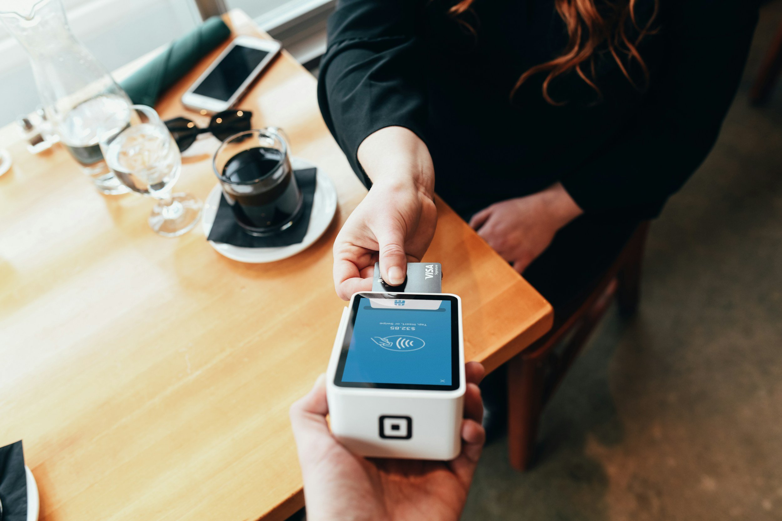 Person making a contactless payment using a mobile card reader and a credit card, with a early transition to table with drinks and a smartphone in the background.