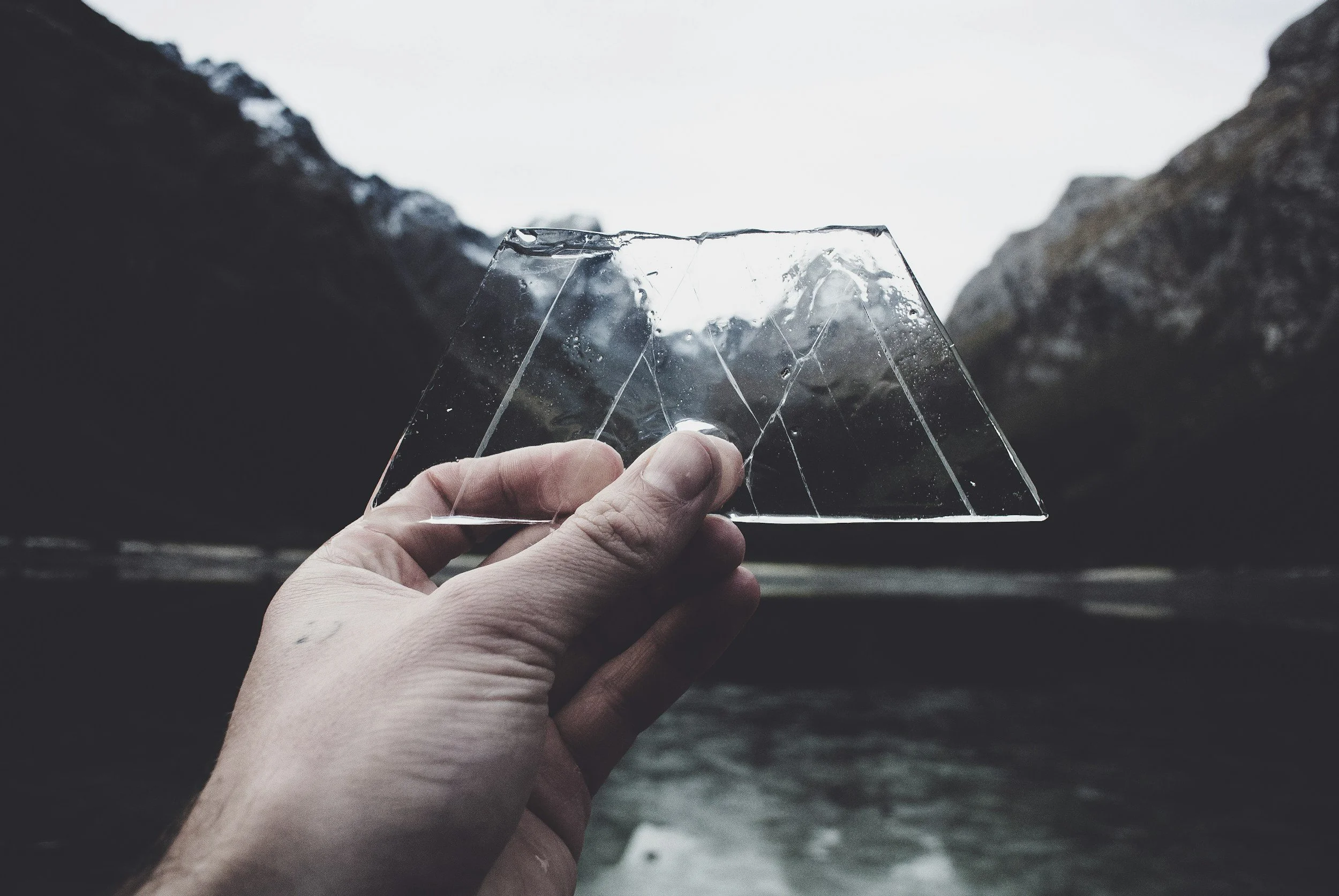 A hand holding a broken piece of glass with a landscape of rocks and water in the background.