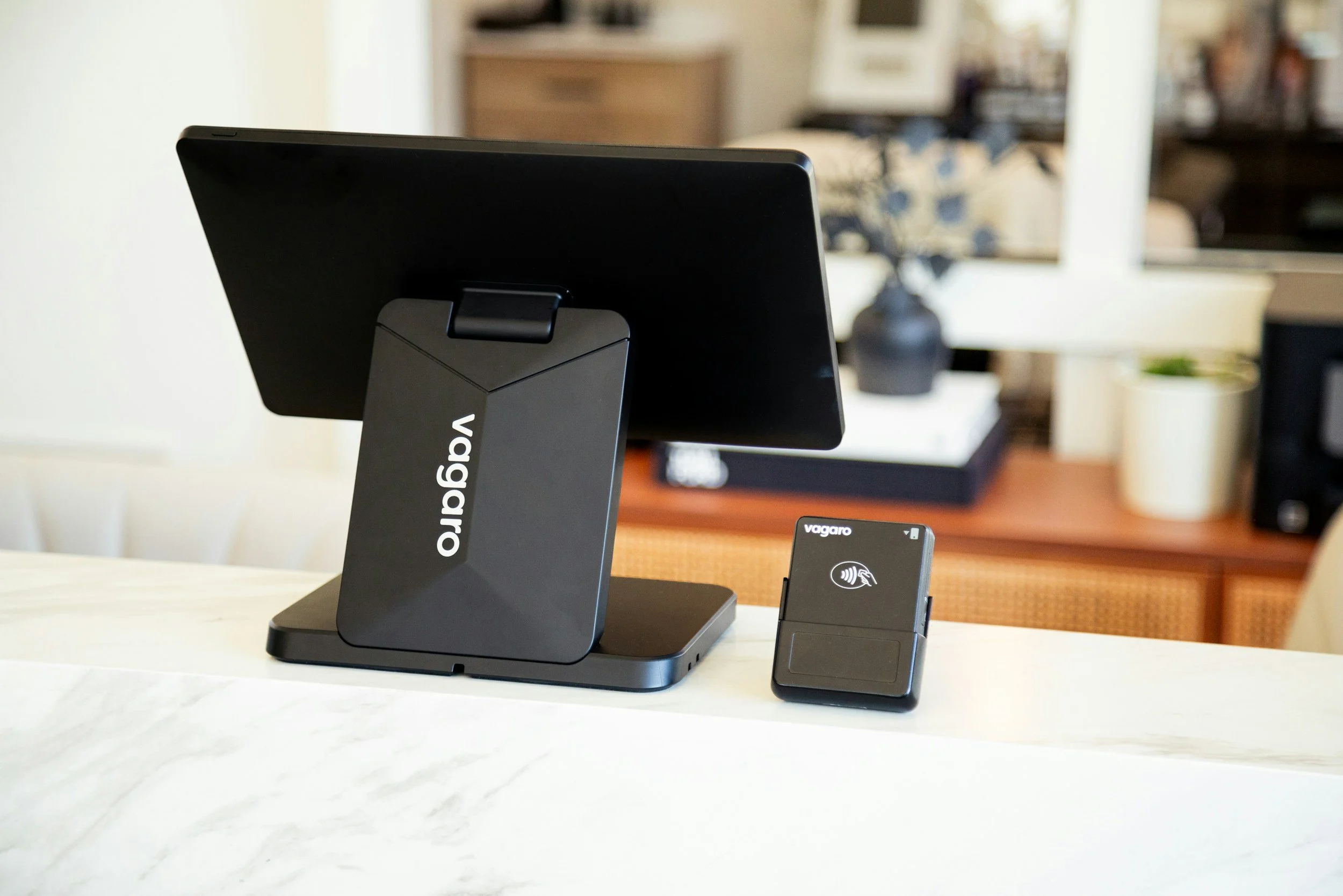 A point of sale (POS) system with a touchscreen monitor and a contactless payment terminal on a white countertop in a retail environment.