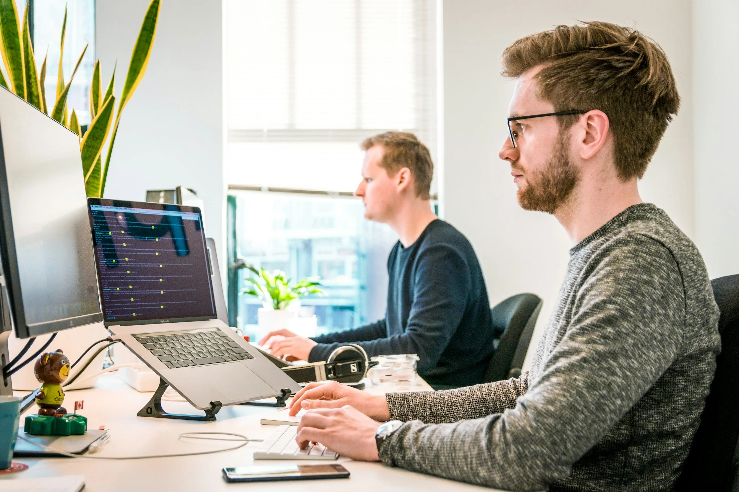 Two men working at desks with computers in an office, one in the foreground using a keyboard and the other in the background typing on a laptop.
