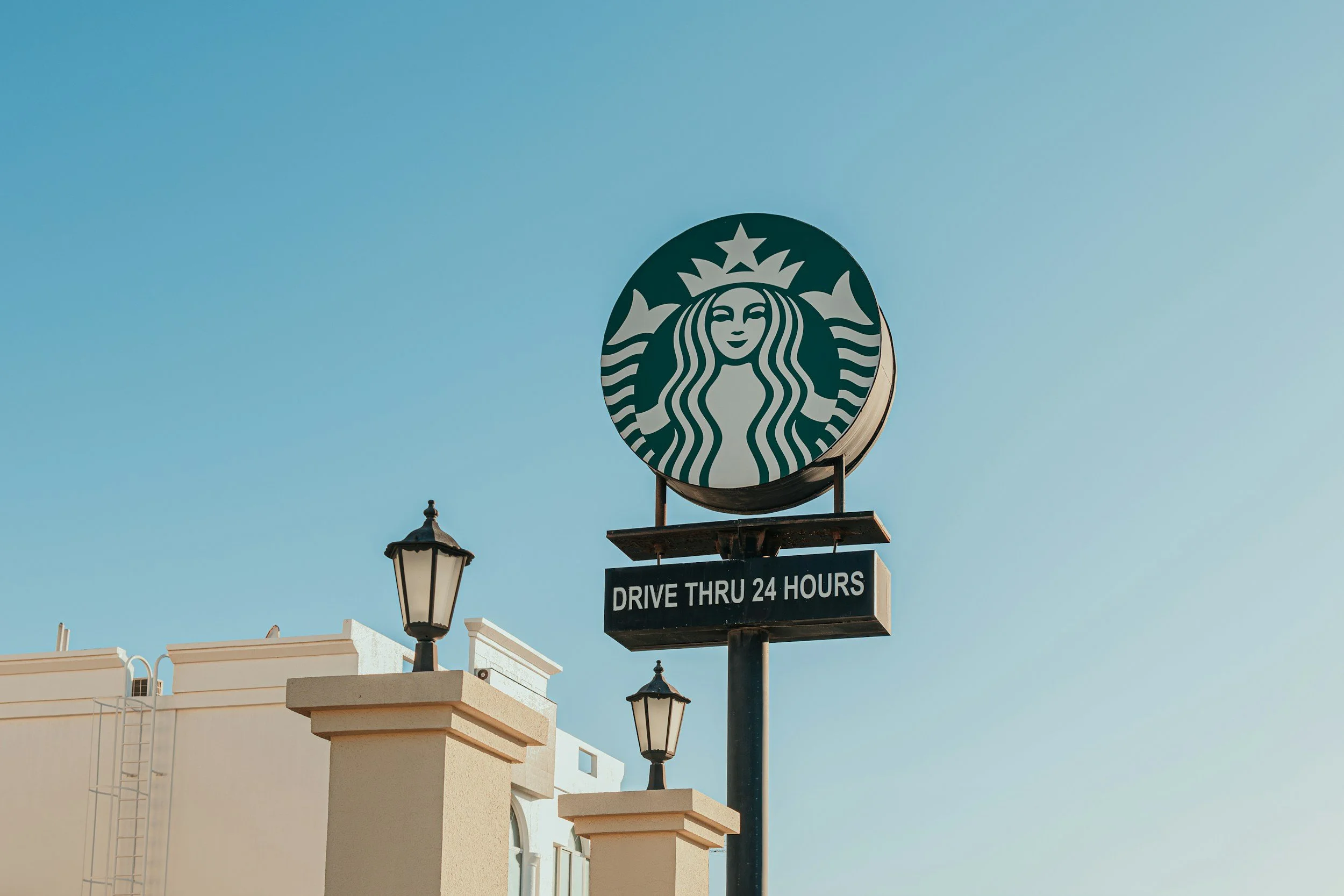 Starbucks logo on a sign with a message saying drive thru open 24 hours, against a clear blue sky.