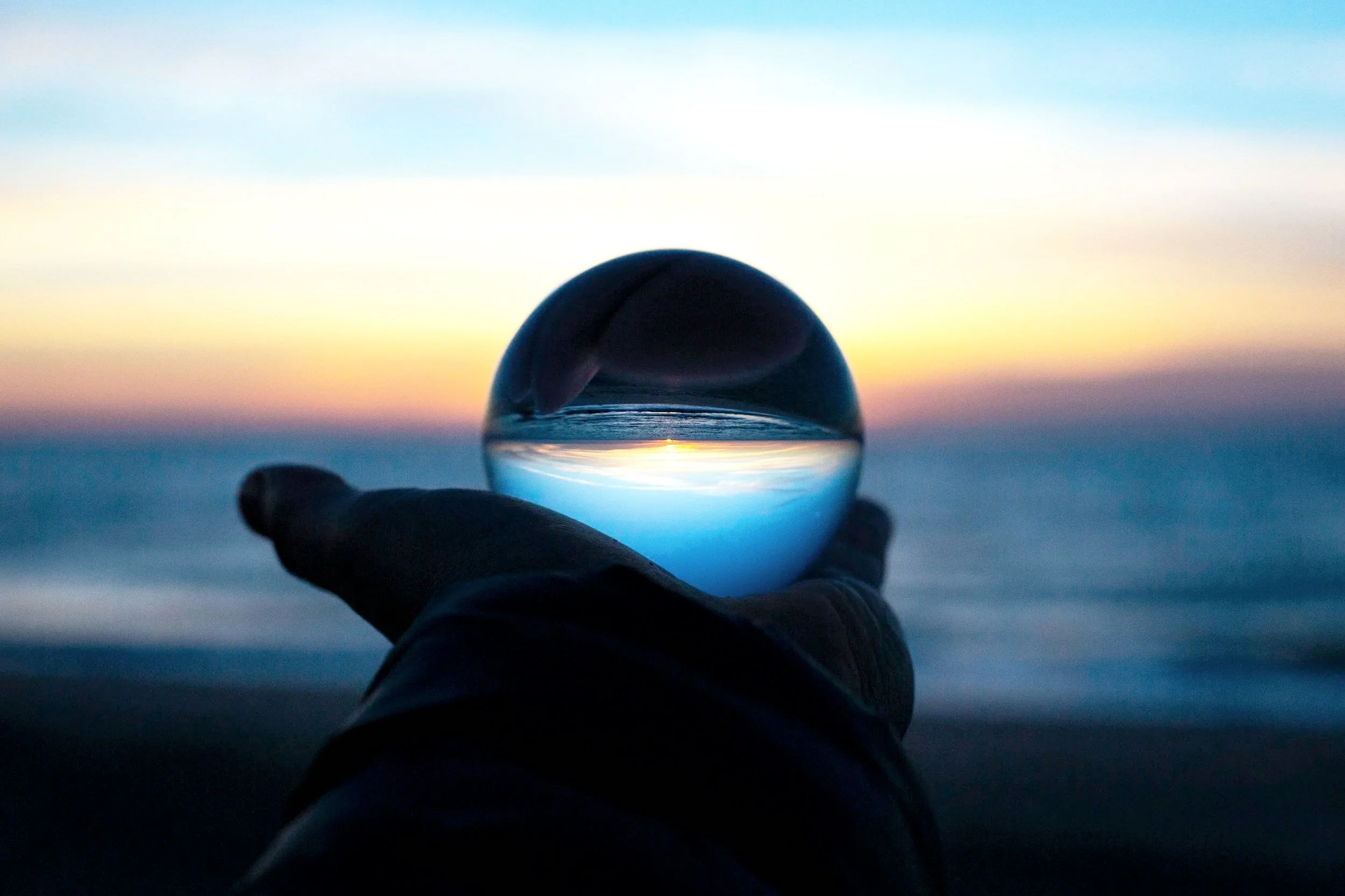 A hand holding a glass sphere on a beach at sunset, with the sky in shades of blue, pink, and orange, and the ocean waves in the background.