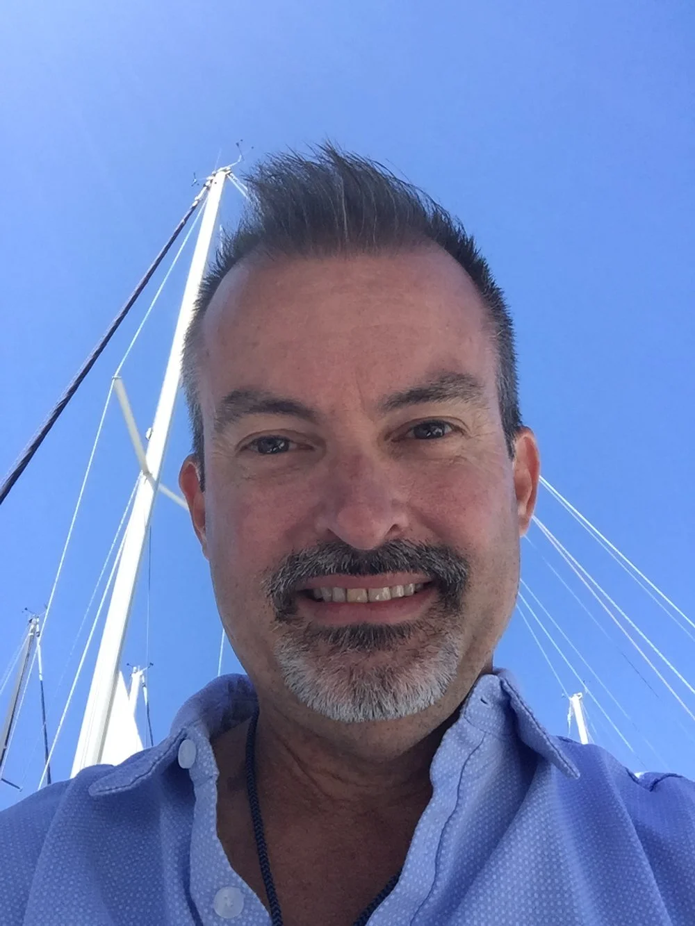 A man taking a selfie outdoors on a clear, sunny day with a blue sky. He has short dark hair, a goatee, and is smiling. In the background, part of a sailboat's mast and rigging are visible.