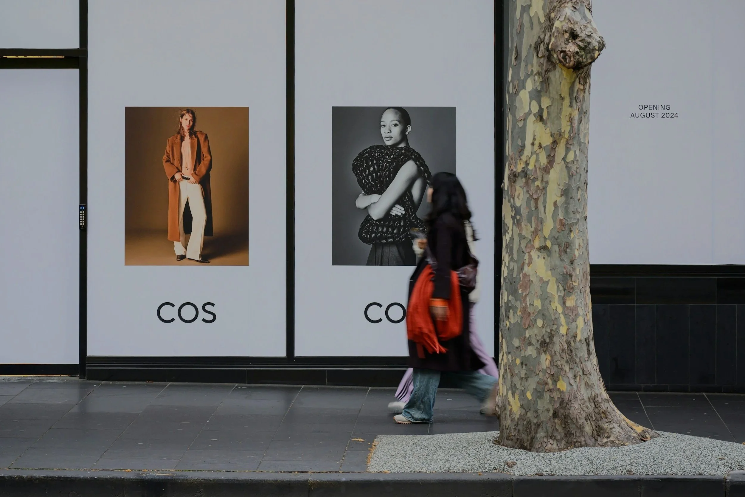 A person walking past a storefront with large posters of fashion models and the text 'cos'. The person is wearing a blackout coat and carrying an orange bag, with a large tree in front of the store.
