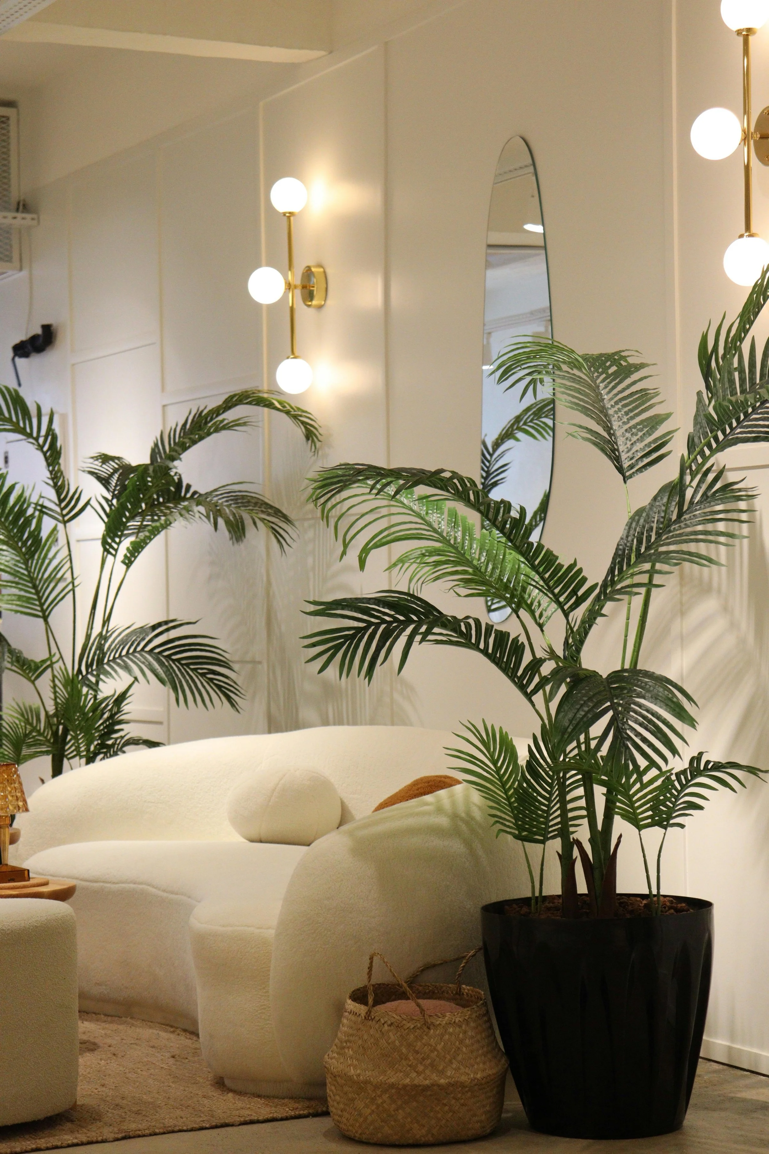 Interior of a living room featuring a white sofa, large potted palm plants, a wall mirror, and modern wall-mounted light fixtures with globe bulbs.