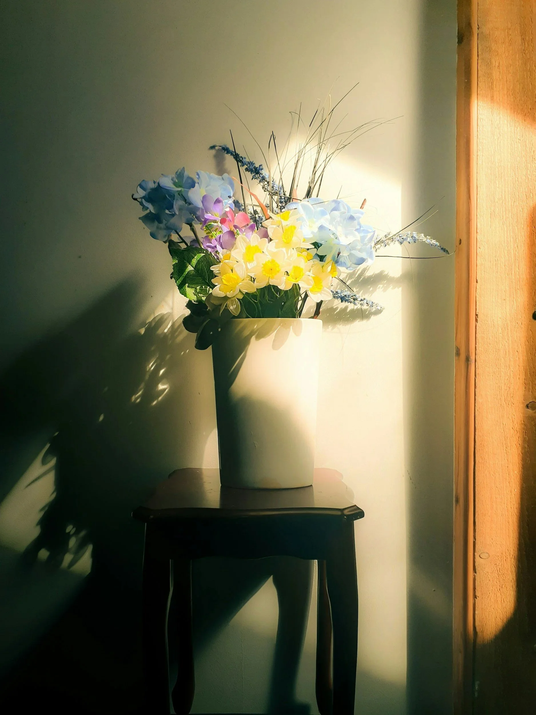 A vase with colorful flowers, including blue, purple, yellow, and white, placed on a small dark wooden table near a wall with warm sunlight casting shadows.