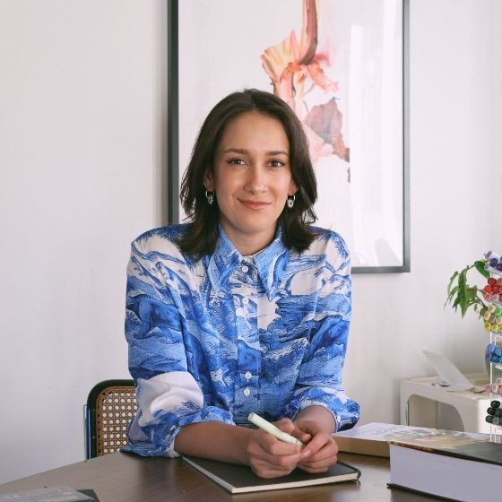 A woman with dark hair wearing a blue patterned shirt sitting at a desk in an office or studio with a artwork on the wall and flowers on the side.