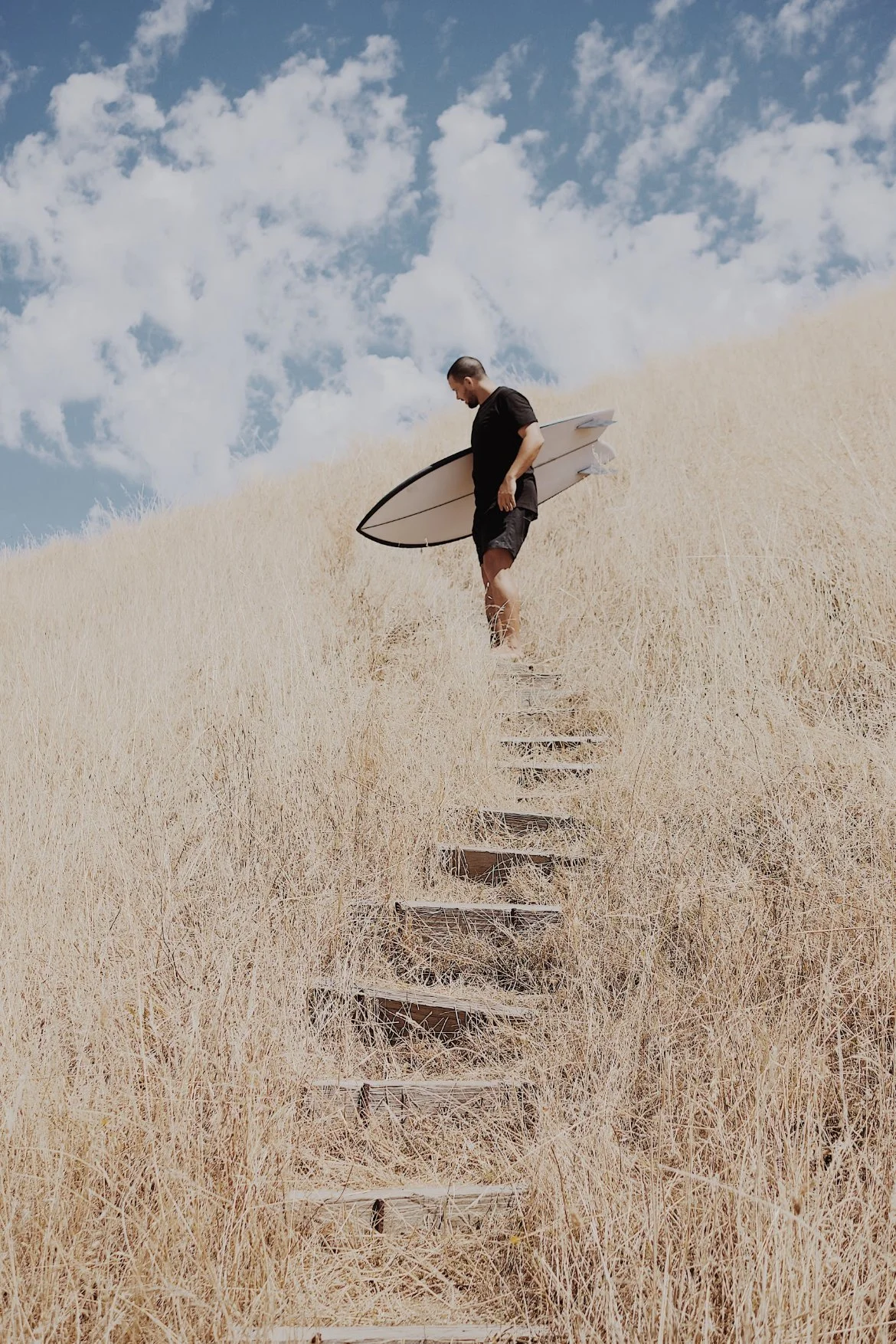 A man holding a surfboard walking down a narrow wooden staircase surrounded by dry grass on a hillside under a partly cloudy sky.