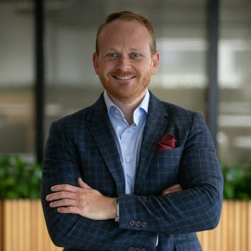 A smiling man in a dark plaid suit with a white shirt, standing with arms crossed in an office or modern building with glass and greenery in the background.