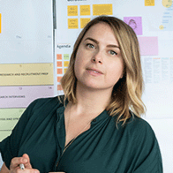 A woman with shoulder-length blonde hair wearing a dark green top, standing in front of a blurred office wall with charts and sticky notes.