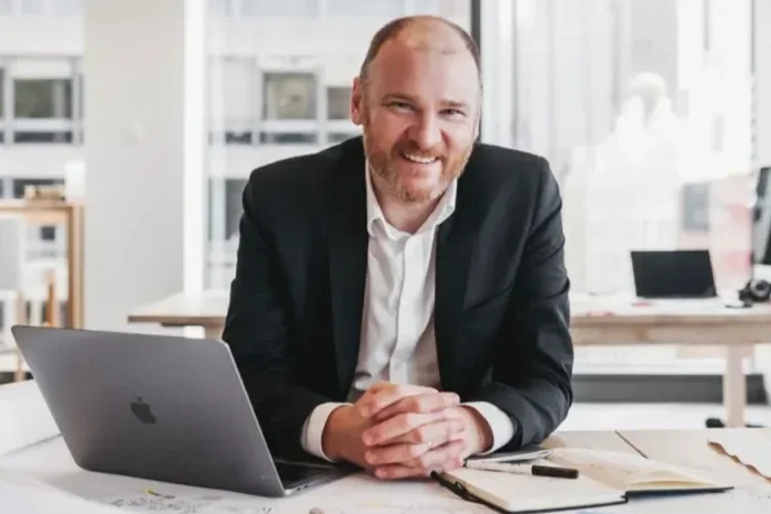 Man in black suit sitting at a desk with open laptop and notebooks, smiling in an office environment.