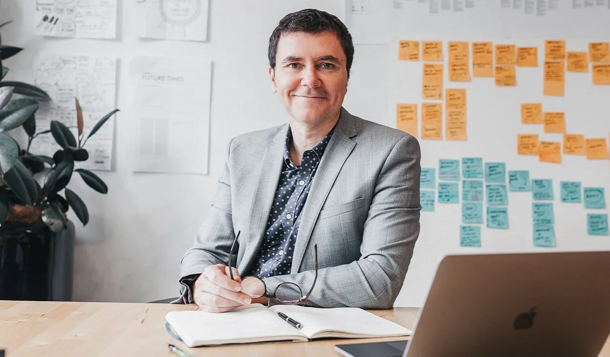 A man in a gray suit sitting at a desk with an open notebook and a pen, in an office with a white wall covered in colorful sticky notes and papers.