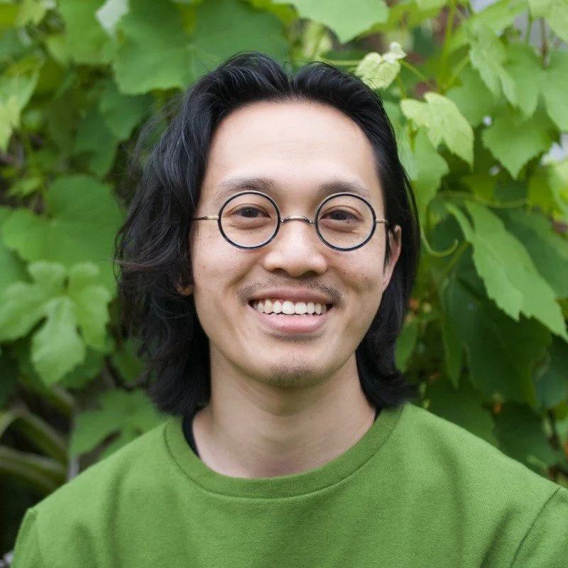 A smiling man with shoulder-length black hair, round glasses, and a green shirt standing outdoors in front of green foliage.