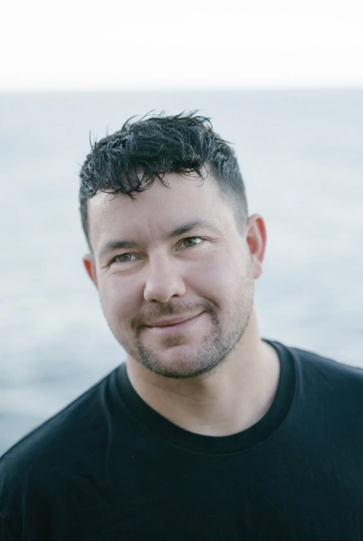 Portrait of a man with dark, curly hair and light facial hair, smiling slightly at the camera, with a blurred body of water in the background.