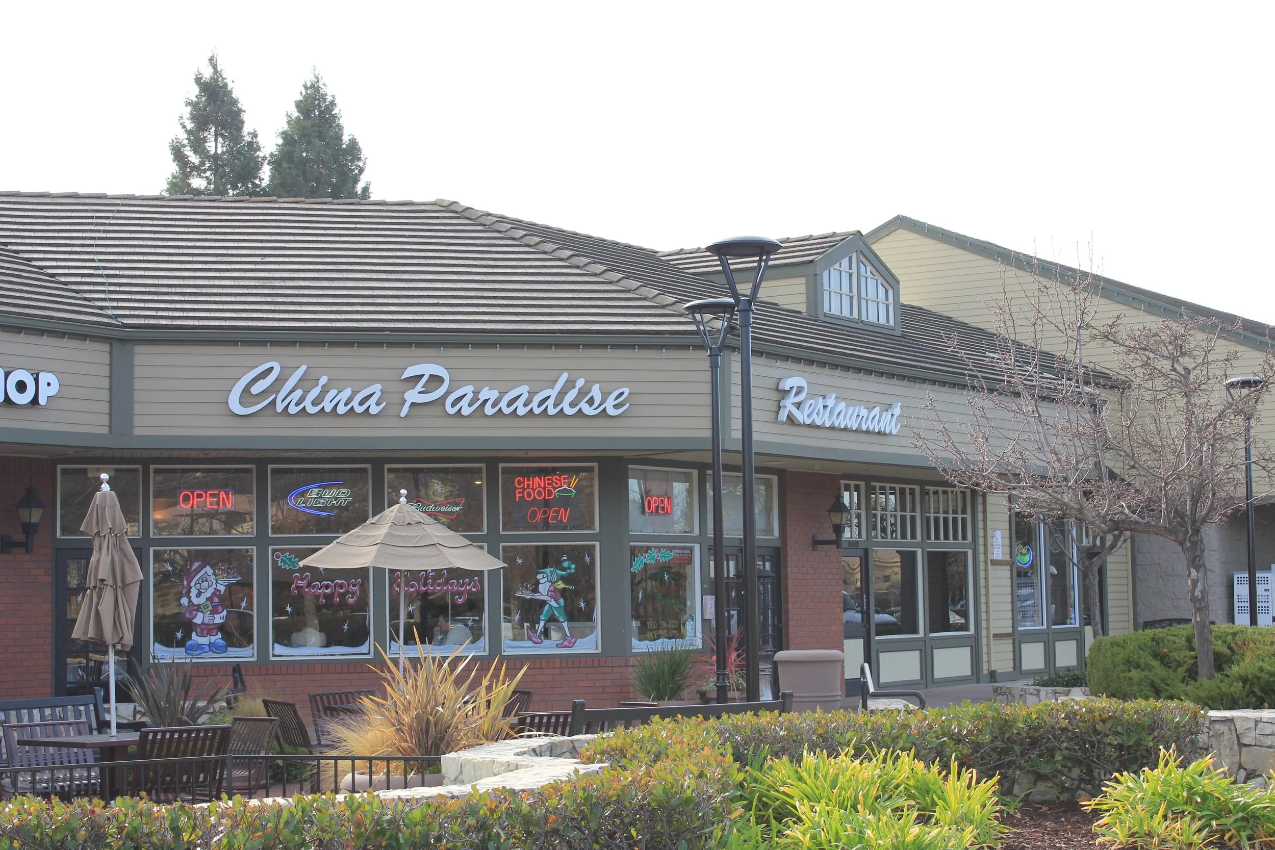 Exterior of a restaurant named China Paradise with Christmas decorations in the windows, outdoor seating area with a closed umbrella, trees, and landscaping in front.