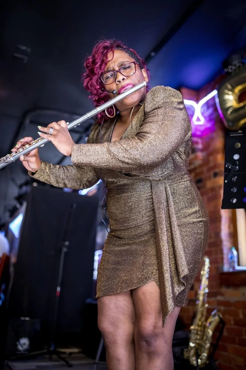 A woman with short, curly, burgundy hair and glasses, wearing a shimmering gold dress, plays a silver flute passionately in a dimly lit venue with neon lights and a brick wall background.
