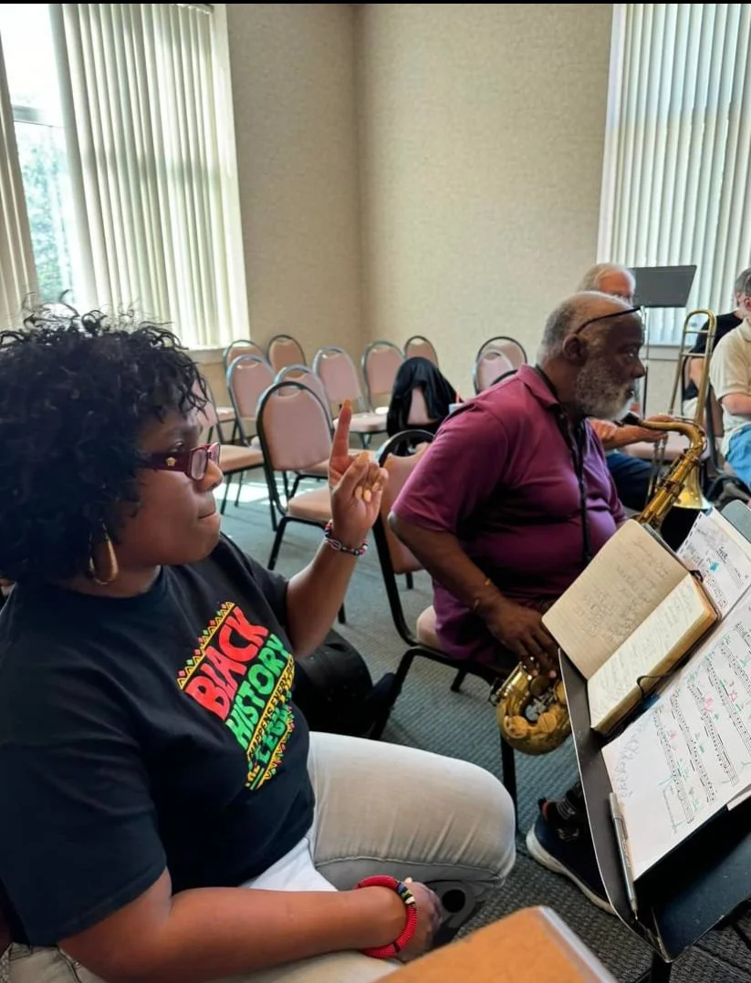 A woman with curly hair and glasses, wearing a Black History shirt, sitting in a music classroom. She is holding up her finger as if making a point, with a music stand in front of her holding sheet music. Behind her, an older man with gray hair and a