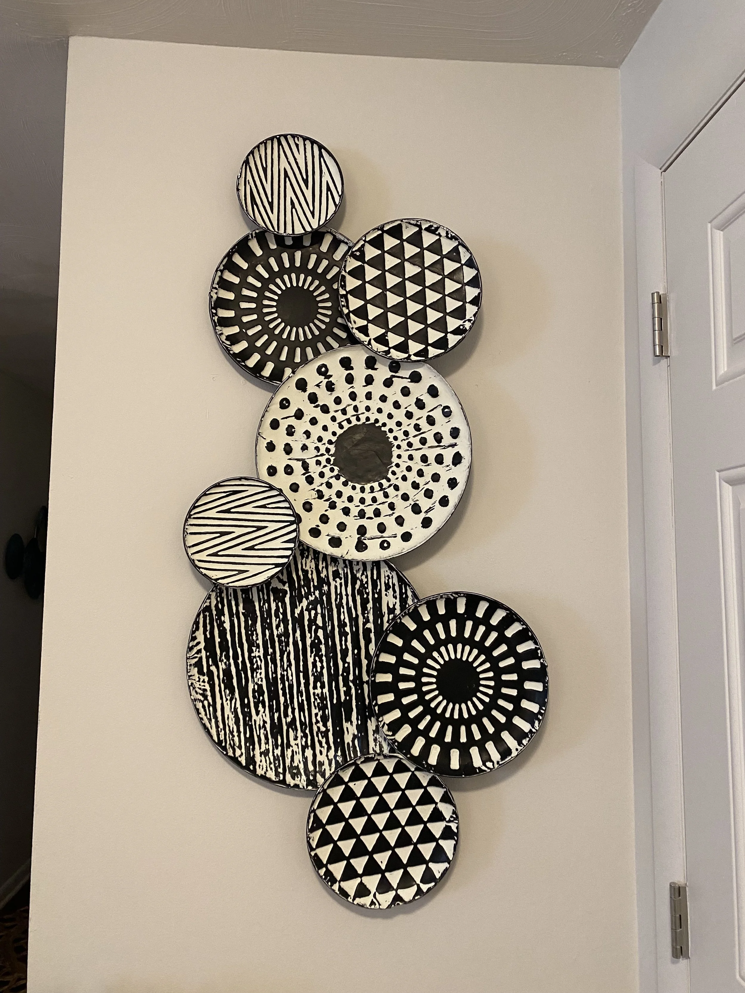 A wall hanging of nine black and white patterned decorative plates arranged vertically on a white wall.