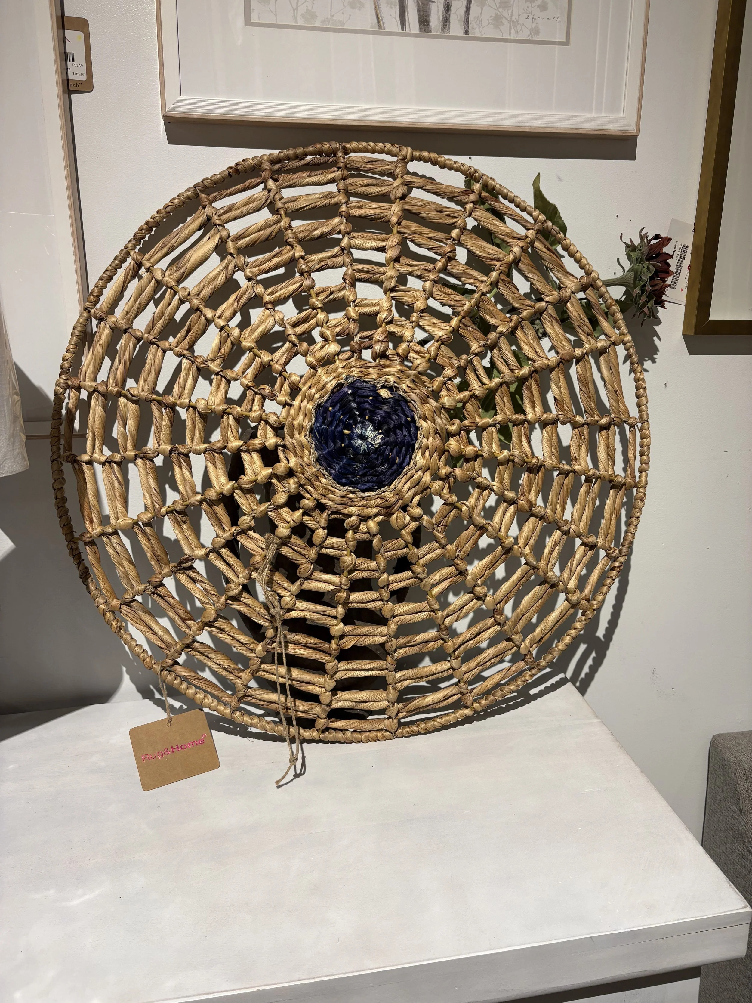 A round woven basket or hat with an open spiral design, made of natural and dark fibers, placed on a white surface indoors.