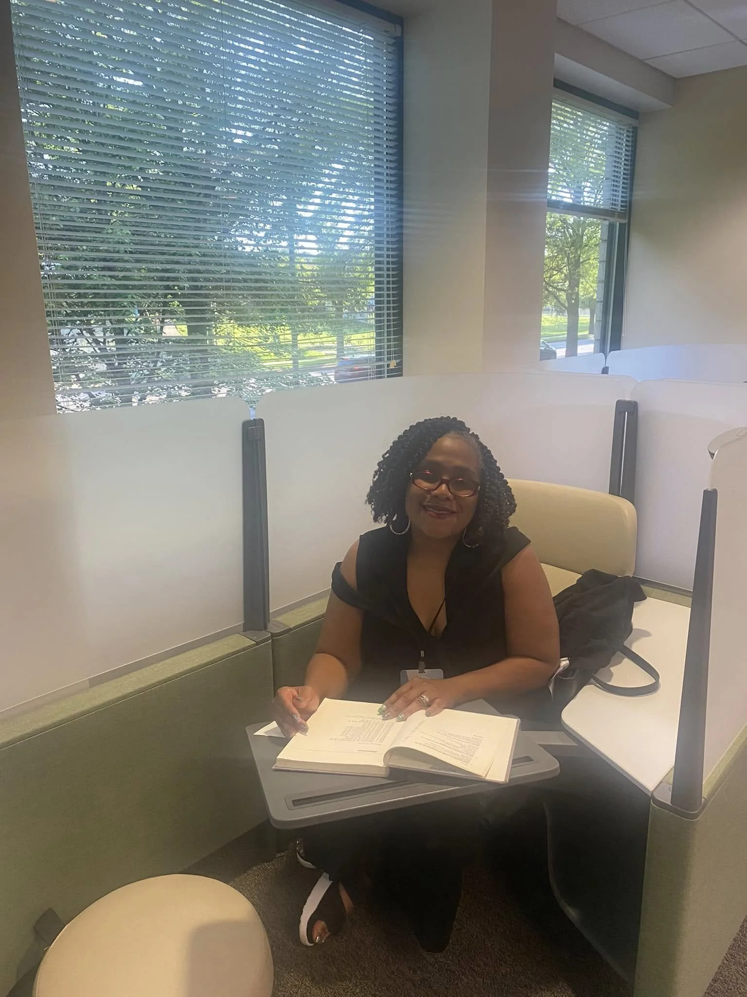 A woman with curly black hair, glasses, and hoop earrings, sitting in a cubicle with a beige chair, reading a book on a lap desk, with windows showing trees and a parking lot outside.