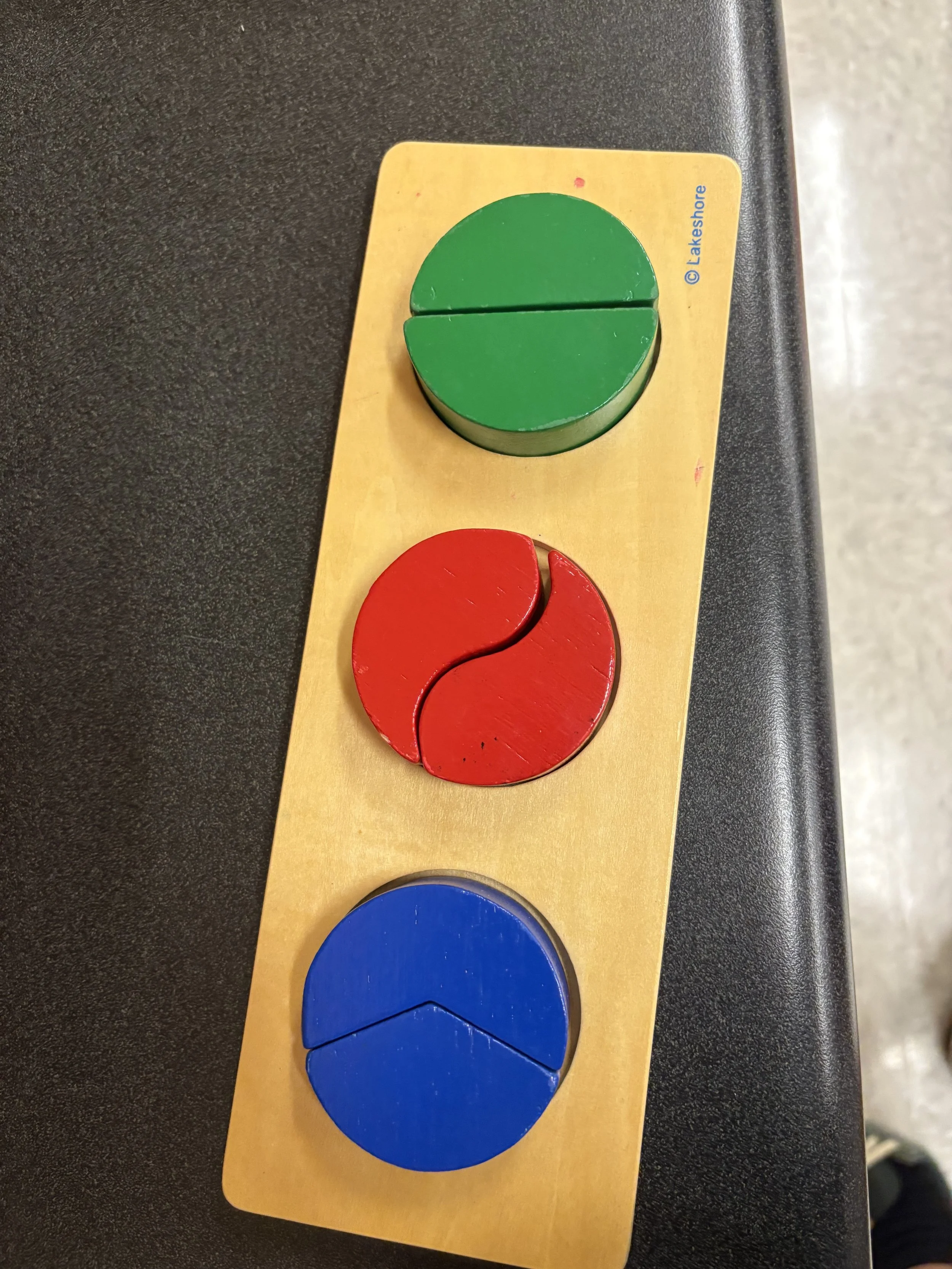 Three round, colored wooden tiles in green, red, and blue mounted on a yellow rectangular board, used for controlled, rhythmic movements.