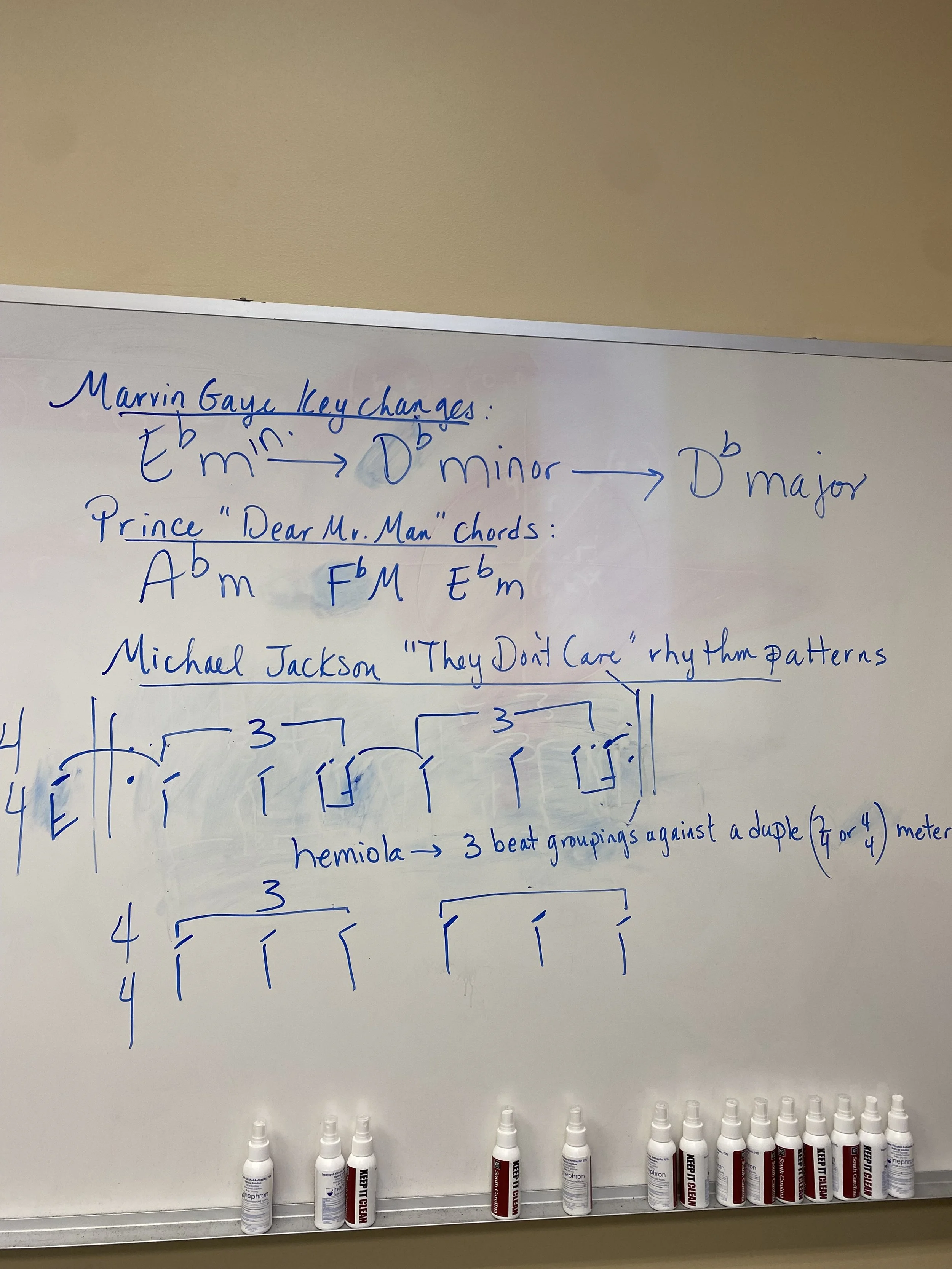 Whiteboard with musical notes, chords, and rhythm patterns written in blue marker, and several white correction fluid bottles placed on the bottom of the board.