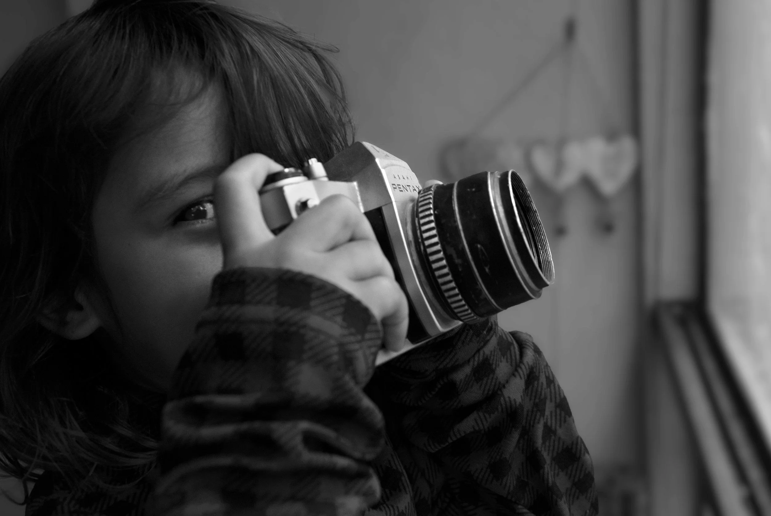 A child with dark hair looks through a vintage film camera, with a focus on the camera and the child's face partially visible, indoors near a window.