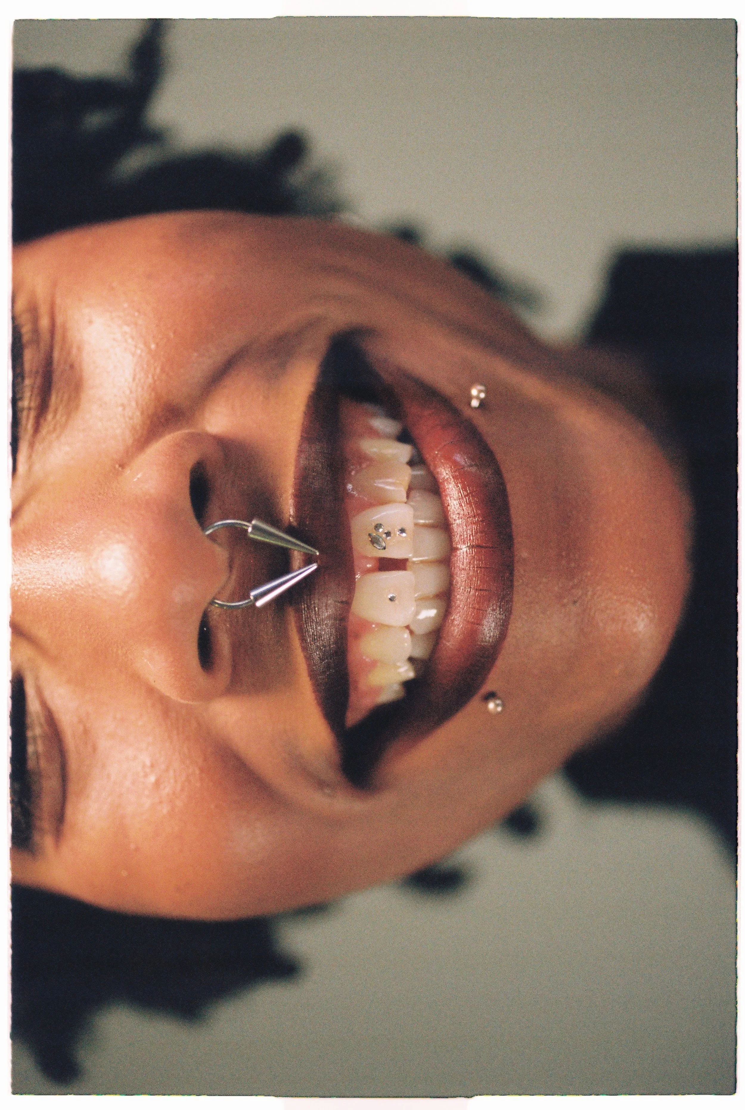 Close-up of a person's mouth with gold lipstick, gold facial piercings, and teeth with jewelry, featuring gold and jewel decorations on the teeth.