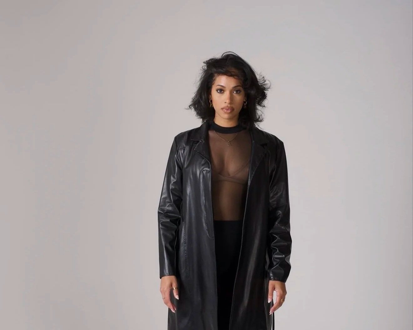 A woman with dark, curly hair wearing a black leather jacket over a sheer black top, standing against a plain light gray background.