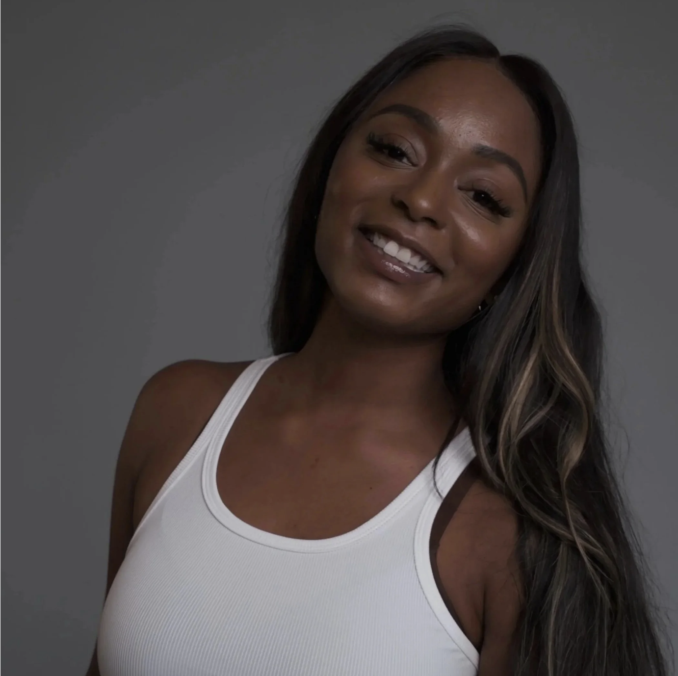 A young woman with long, wavy dark hair and brown highlights, smiling and wearing a white tank top, standing against a gray background. She is Rashauna Payne the Founder of MuseWorks.