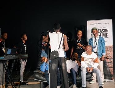 Group of people on stage during a rehearsal or event, some holding scripts or microphones, with a piano on the left and a banner in the background.