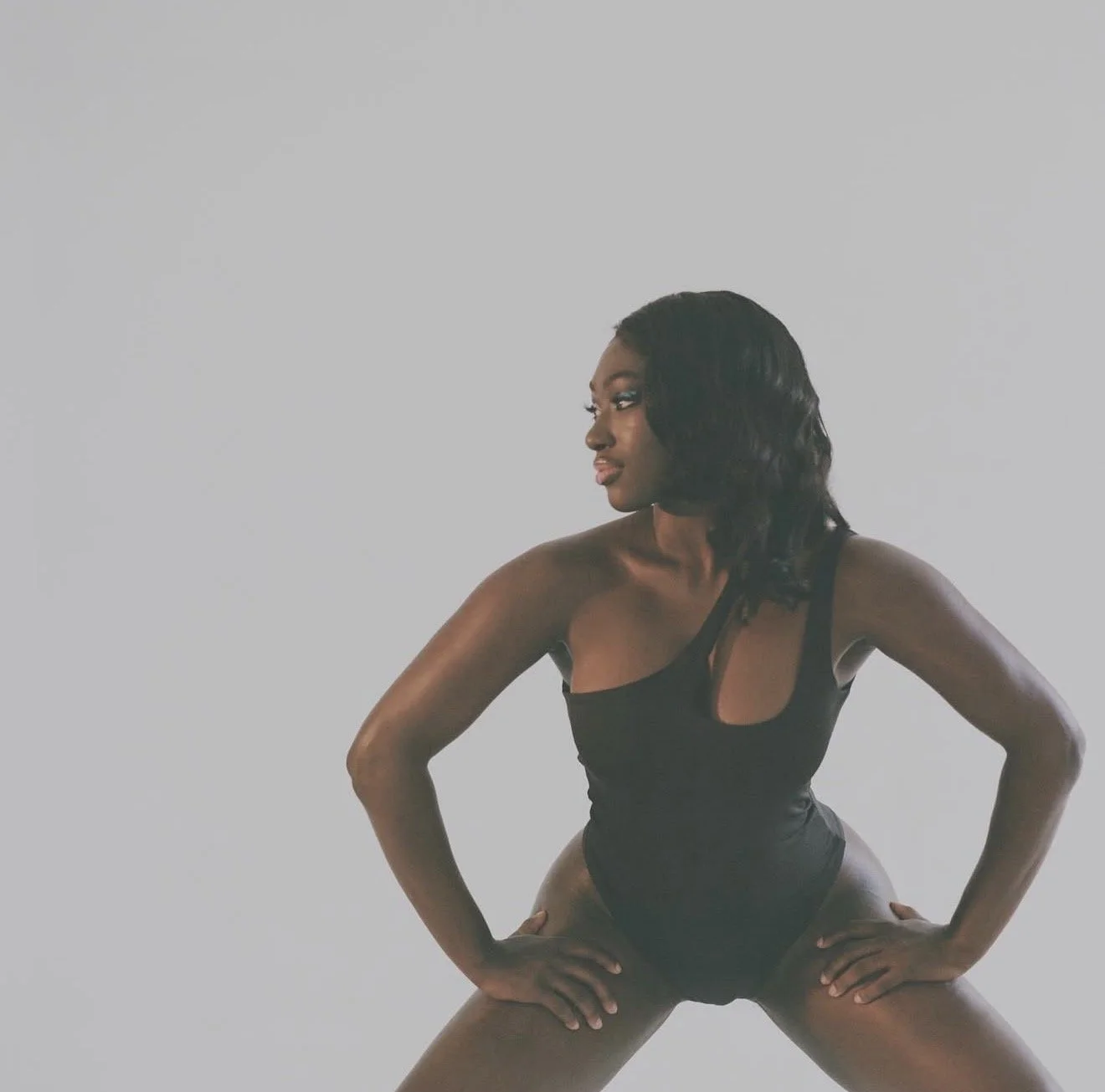 A woman with dark hair in loose waves, wearing a black sleeveless bodysuit, standing with legs apart and hands on hips, against a plain light gray background.