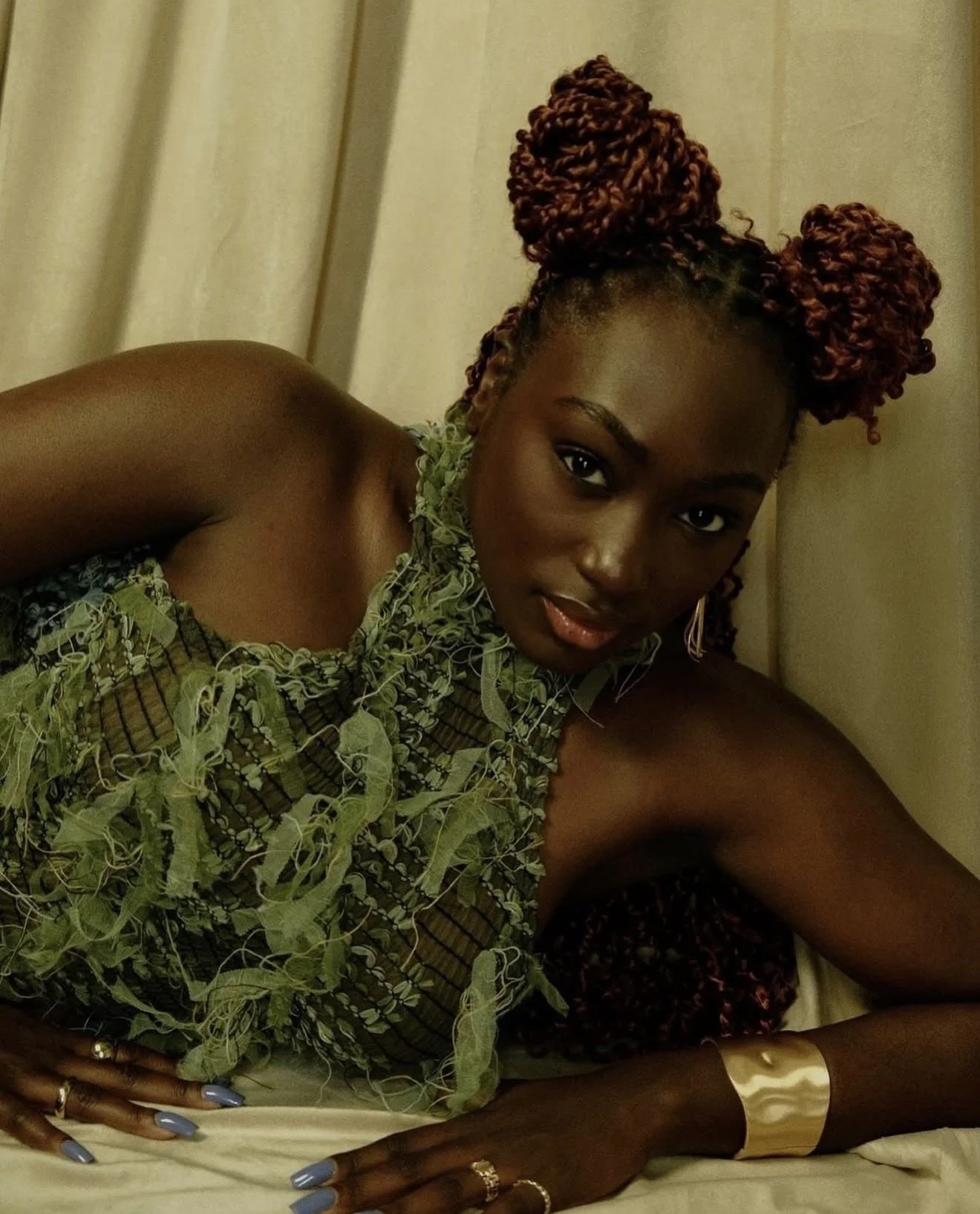 A woman with dark skin, styled in braided hair with a reddish hue, is lying on her side on a bed with cream-colored bedding. She is wearing a green, seashell-patterned top, and has jewelry including rings, earrings, and a gold cuff bracelet. She looks directly at the camera with a relaxed expression, against a background of cream curtains.