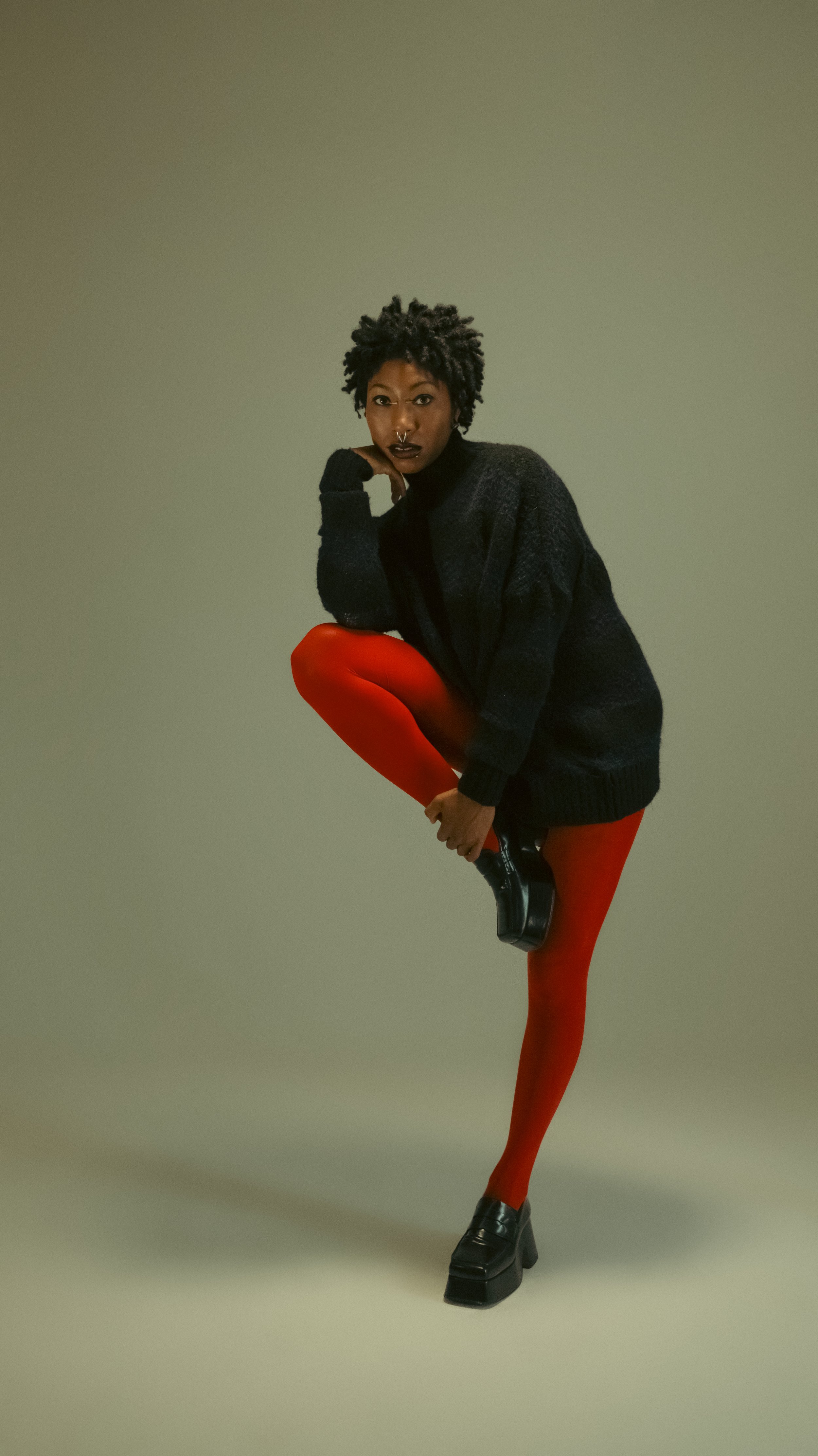 Person with short curly hair wearing a black sweater, red tights, and black shoes, standing on one leg while holding the raised foot with their hand, against a plain background.