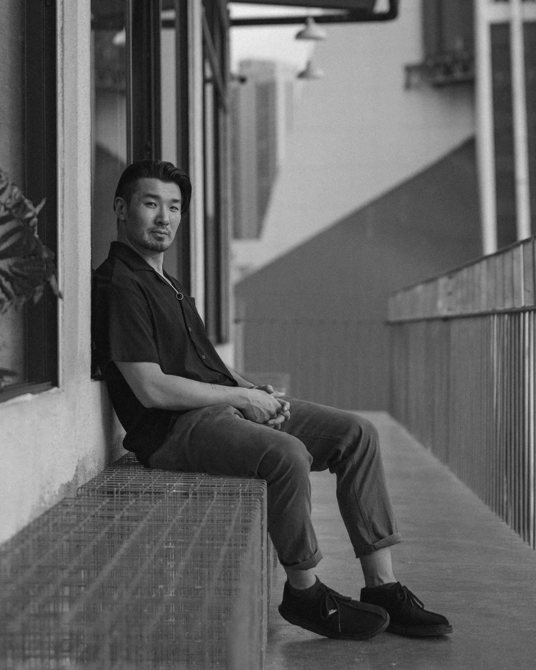 A man sitting on a bench on a balcony, looking at the camera, in black and white.