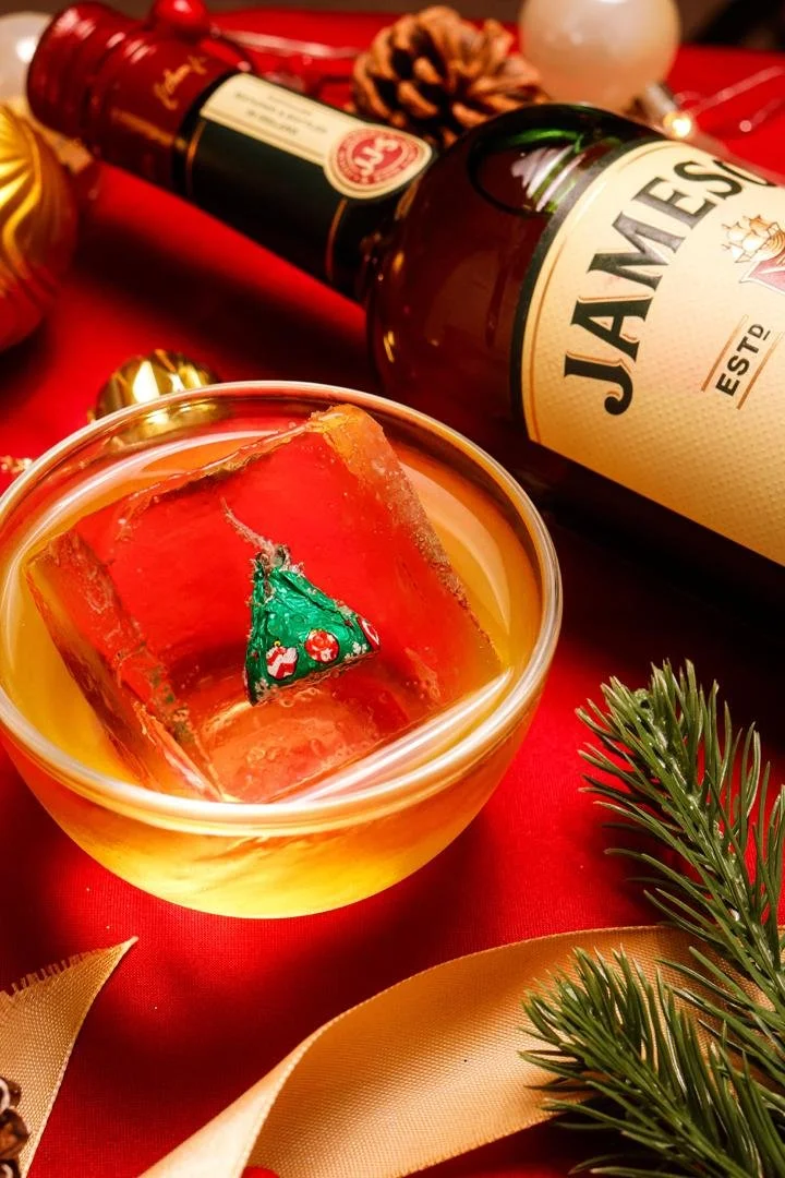 A glass bowl with a holiday-themed candle inside, featuring a Christmas tree design, on a red tablecloth with holiday decorations, a bottle of Jameson Irish whiskey, pine cones, and green pine branches.